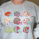 A woman wearing a light gray crewneck sweatshirt with a graphic of nine colorful football helmets on the front, posed in an indoor setting.